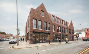 Contemporary brick building with shops at street level and apartments above on a corner urban streetscape, with pedestrians, a cyclist, and parked cars.