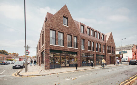 Contemporary brick building with shops at street level and apartments above on a corner urban streetscape, with pedestrians, a cyclist, and parked cars.